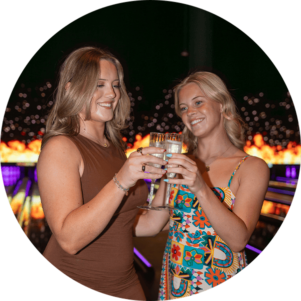 Two women enjoying drinks at a luxury bowling alley with vibrant lighting.